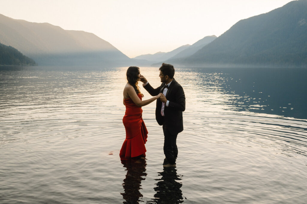 Groom wipes something off the bride's nose while they stand in Lake Crescent