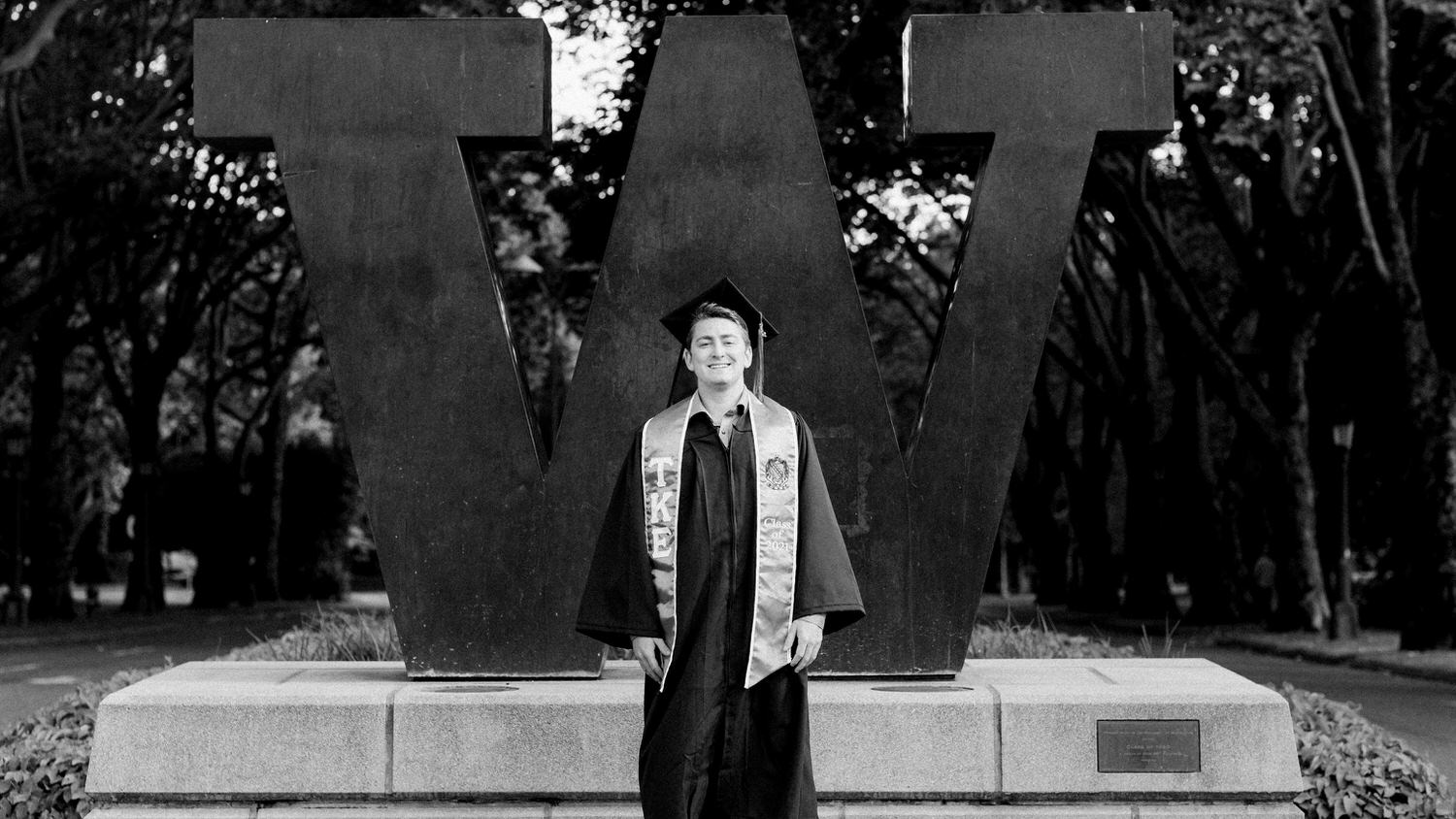 Black and white portrait of University of Washington Graduate standing in front of UW logo in his cap and gown