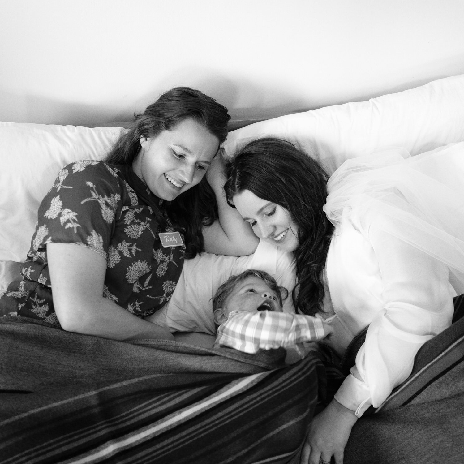 The Best Documentary Wedding Photography of 2025 14 The bride lies on a bed smiling at a newborn surrounded by close friends